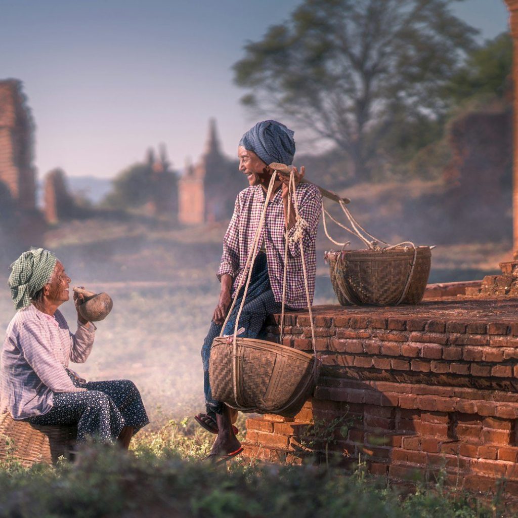 sitting person smiling beside man sitting on brown concrete pavement while carrying baskets on his left shoulder