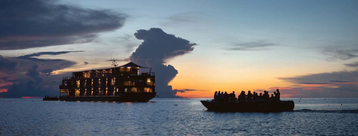 Aqua Mekong lit up at dusk on the Mekong with a tender boat of guests returning from an excursion