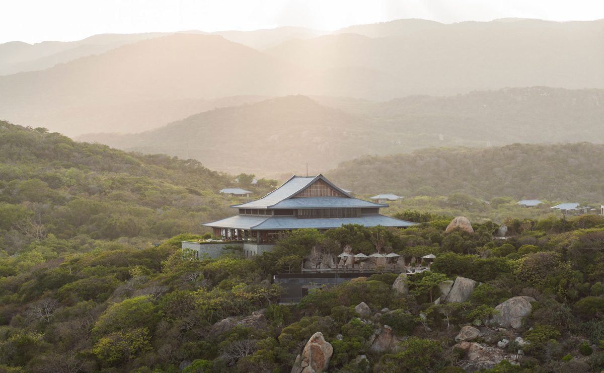 Amanoi's central pavilion with a layered roof on a forested hillside above Vinh Hy Bay, mountains behind